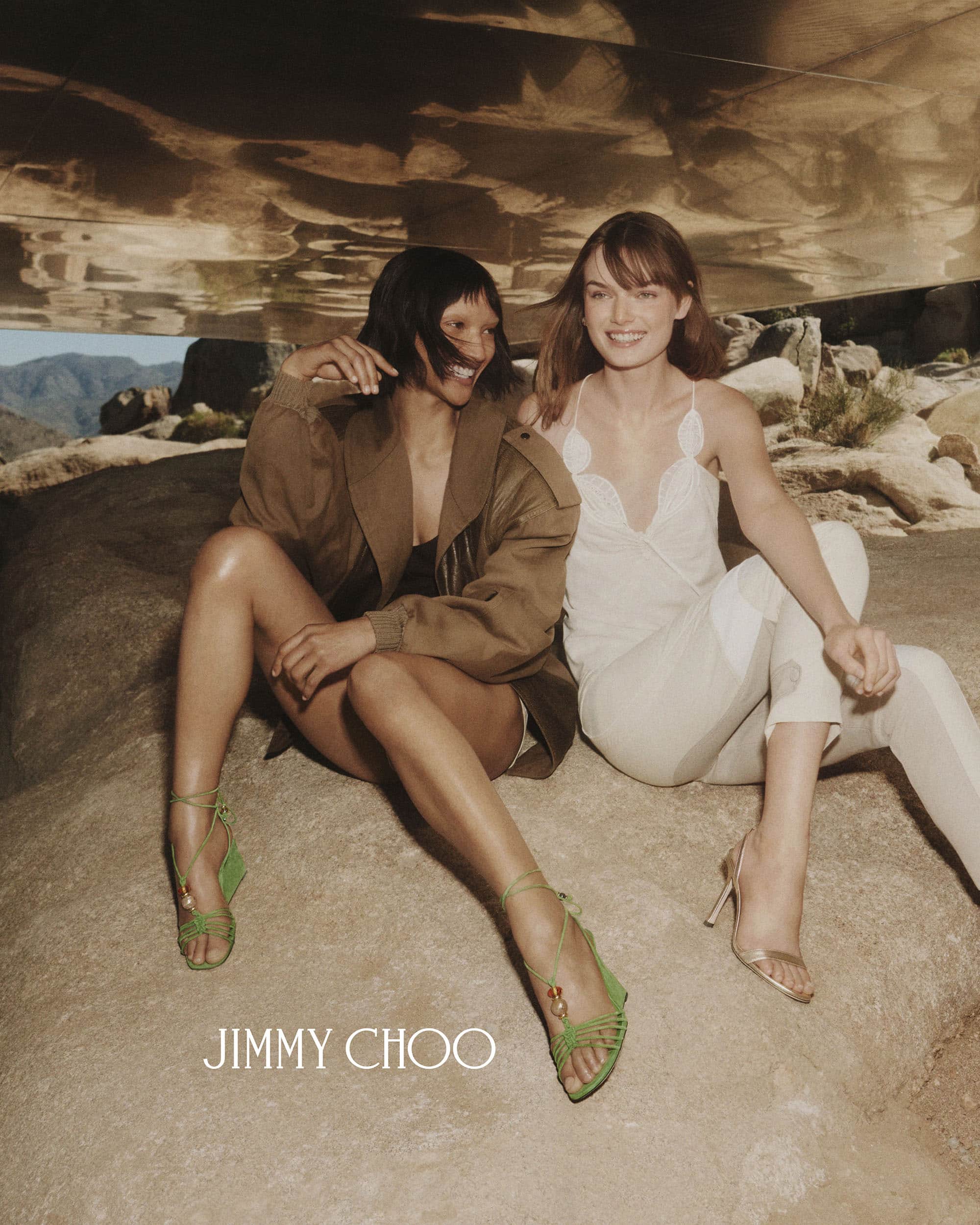 Two female models seated outdoors on rocky terrain wearing designer clothing and strappy sandals for Jimmy Choo ad campaign