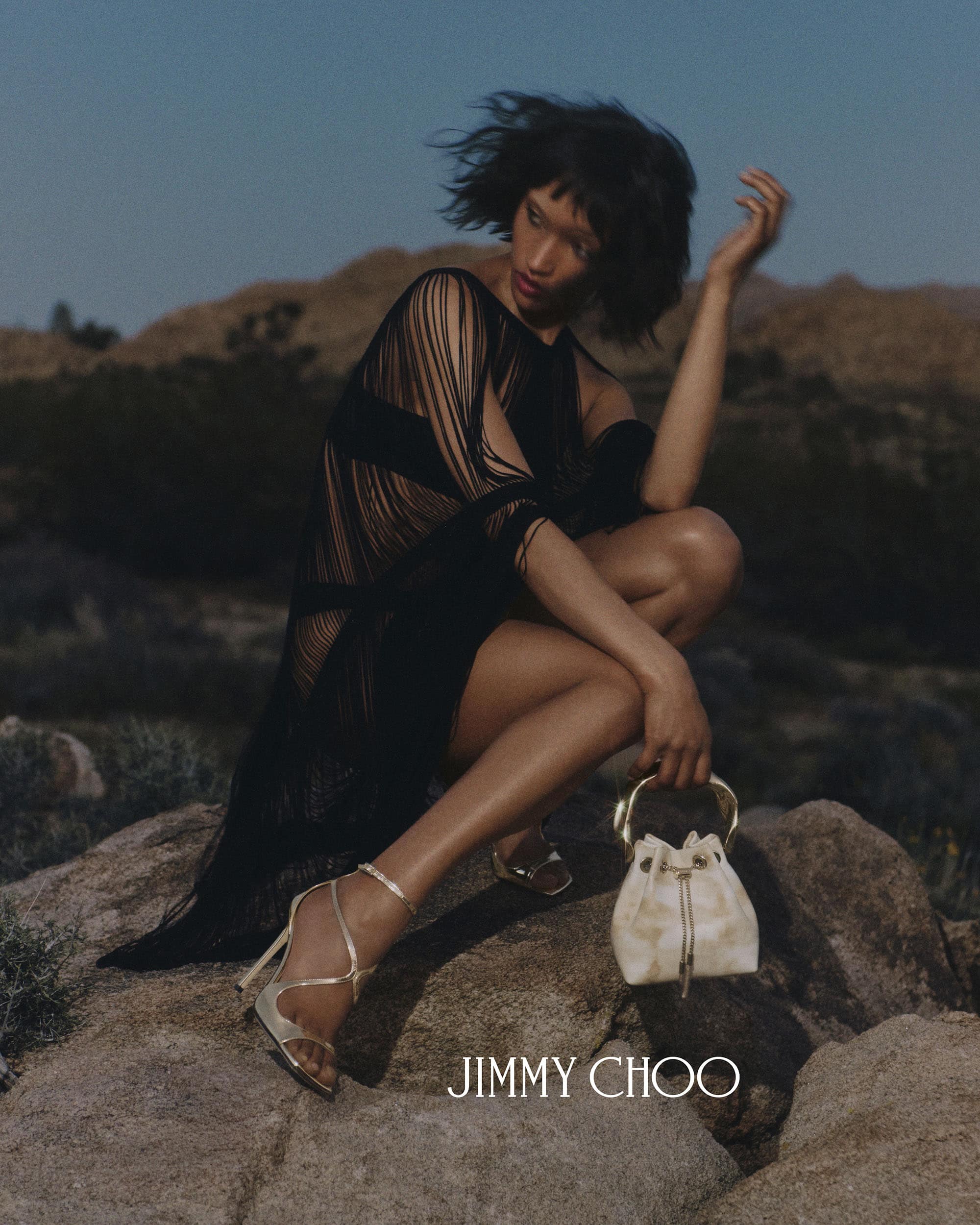 Model in black fringe dress crouched on rocks outdoors holding a small bag for Jimmy Choo summer campaign