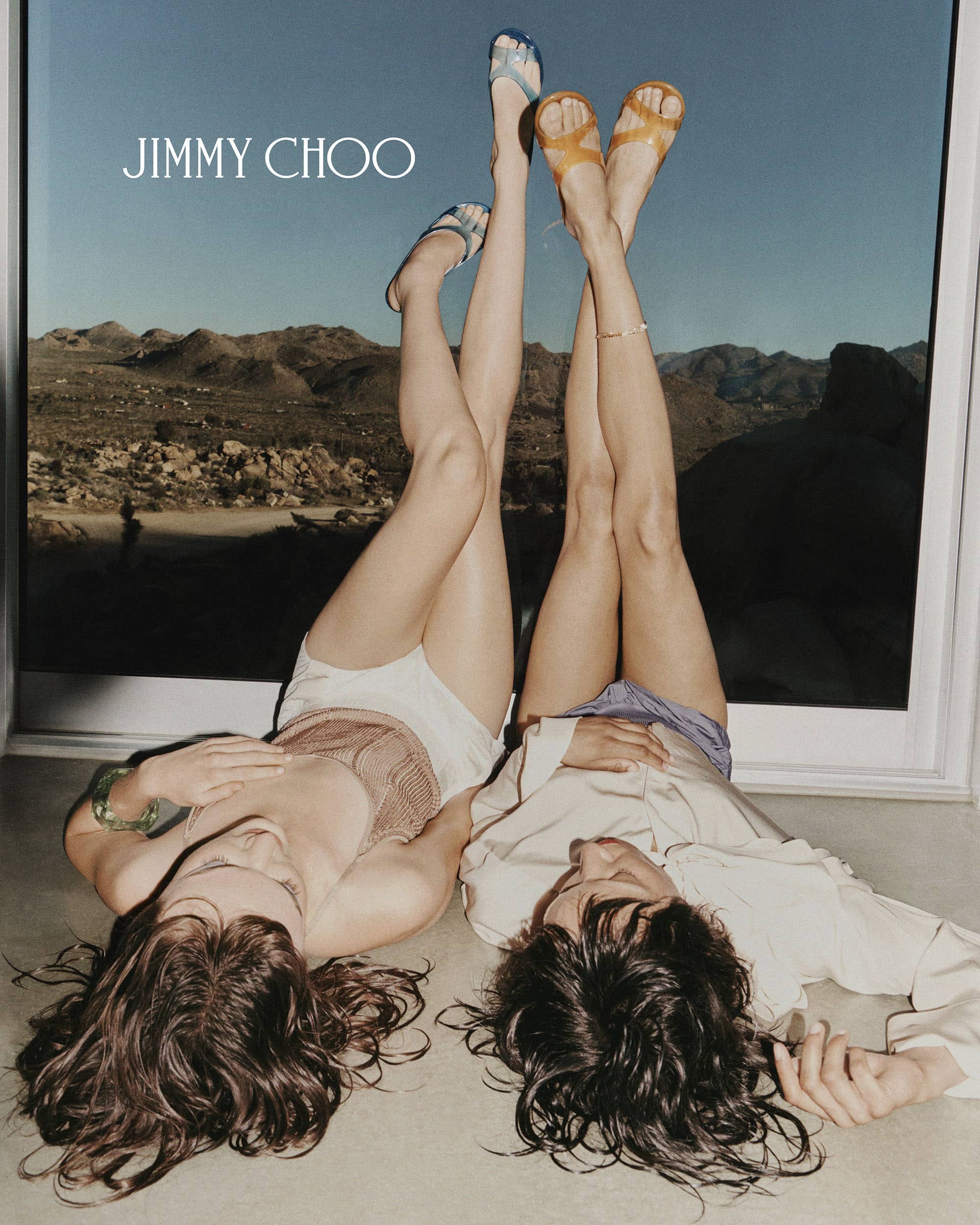 Two women lying on the floor with legs raised against a window showing a desert landscape for Jimmy Choo.