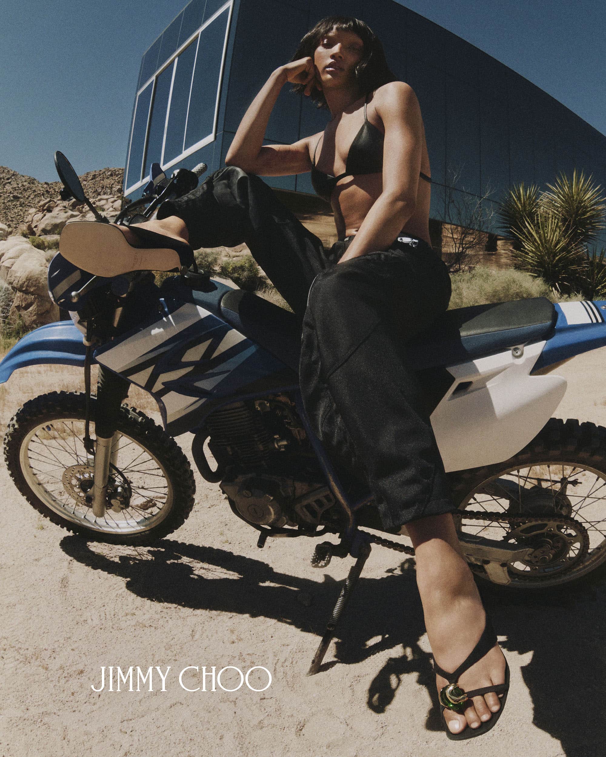 Woman posing on a blue and white dirt bike outdoors wearing black trousers and sandals with a modern building in the background
