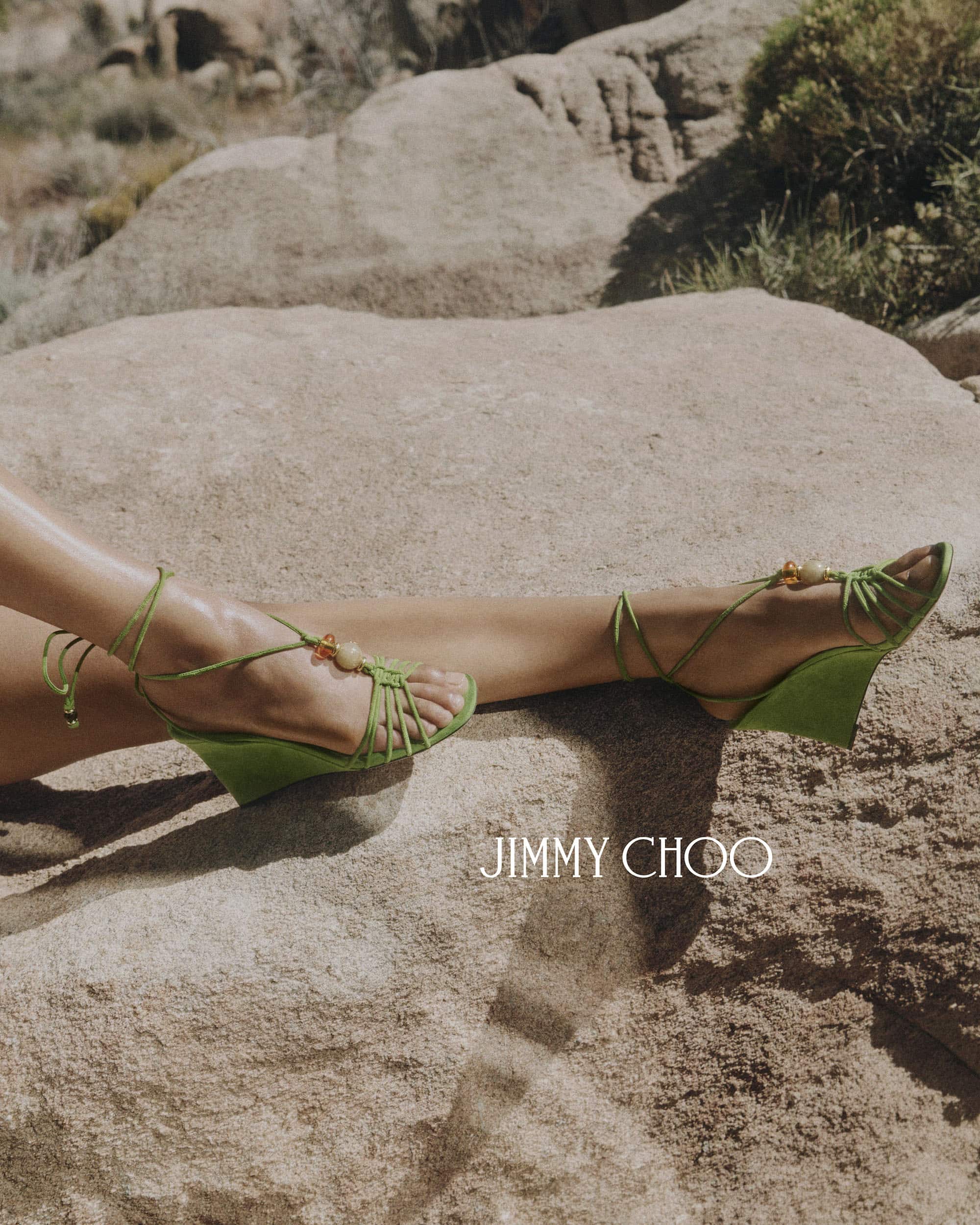 Close-up of legs wearing green strappy wedge sandals resting on natural stone rocks in an outdoor desert landscape.