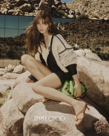 Model crouching on large desert boulders wearing a cream and black jacket while holding a green handbag for Jimmy Choo.