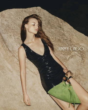 Model in a black sequin dress holding a green suede handbag leaning against a large rock