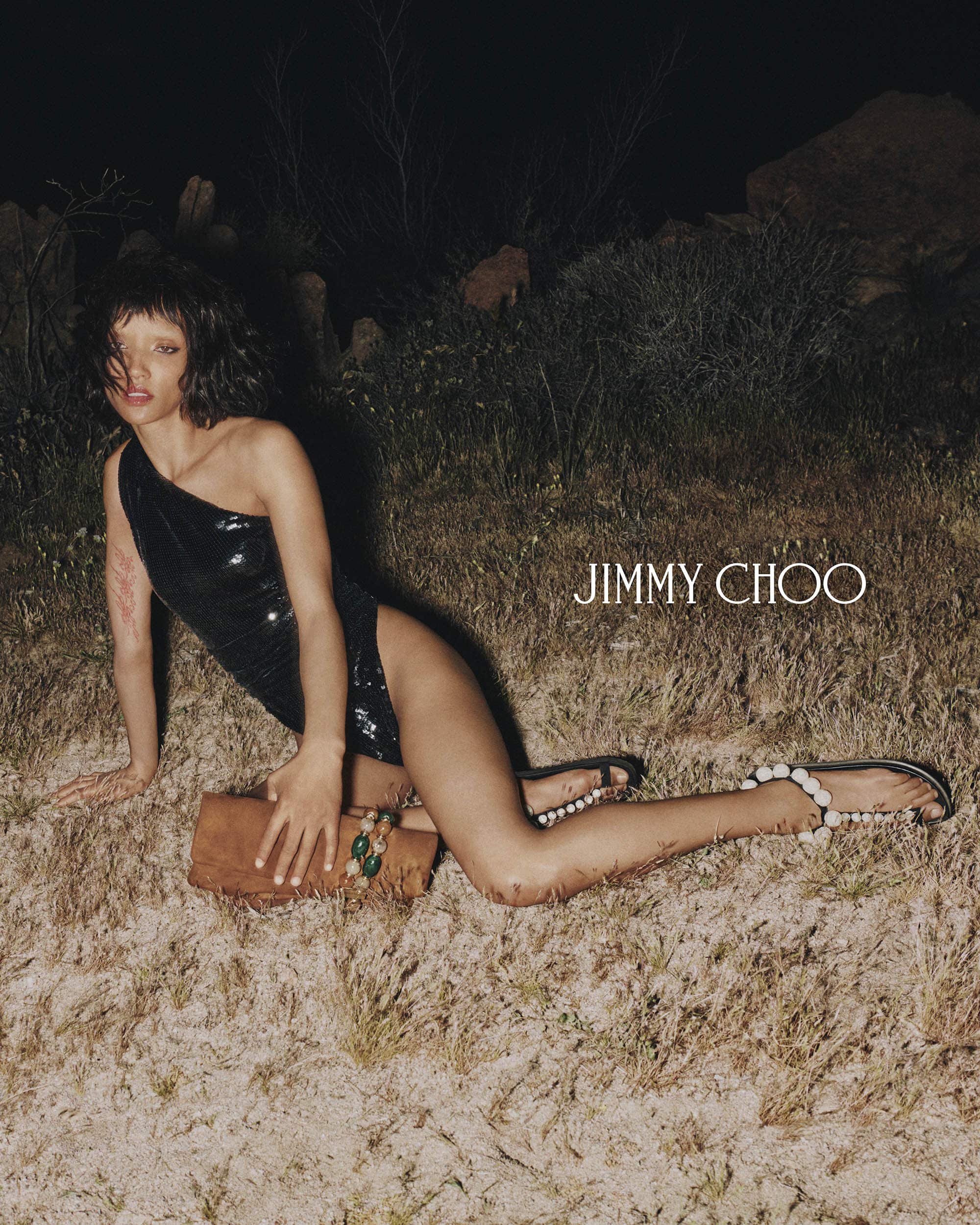 Woman poses in a one-shoulder swimsuit with a tan clutch and black sandals in outdoor fashion shot. Jimmy Choo logo visible.