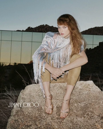 Model wearing a shimmering fringed top and strappy metallic sandals crouches on a desert rock outdoors.