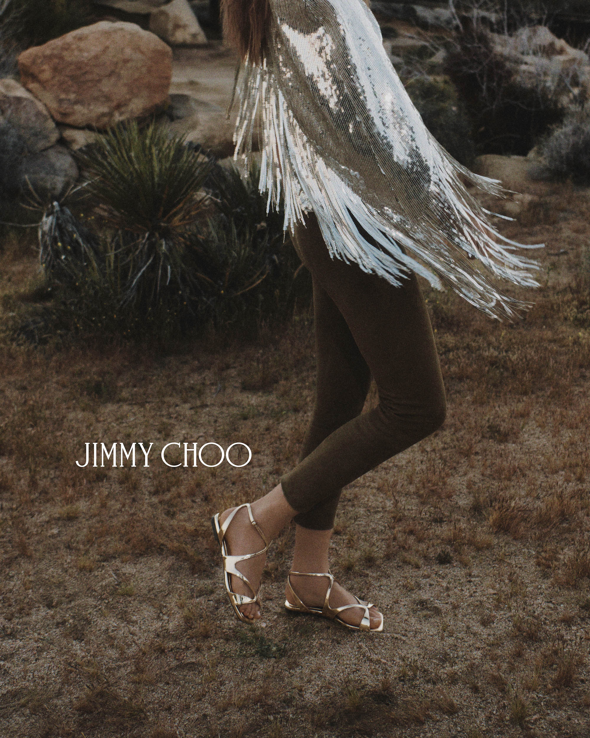Model wearing gold metallic sandals and fringed silver garment standing outdoors near desert vegetation