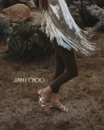Model wearing gold metallic sandals and fringed silver garment standing outdoors near desert vegetation