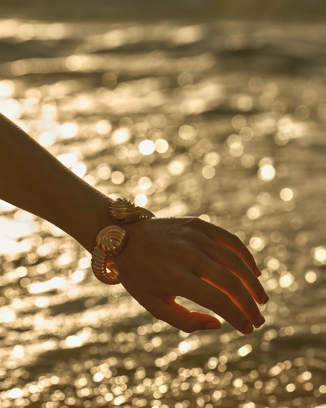 Close-up of a woman's hand adorned with a gold cuff bracelet featuring a spiral design, set against a blurred golden ocean backdrop.