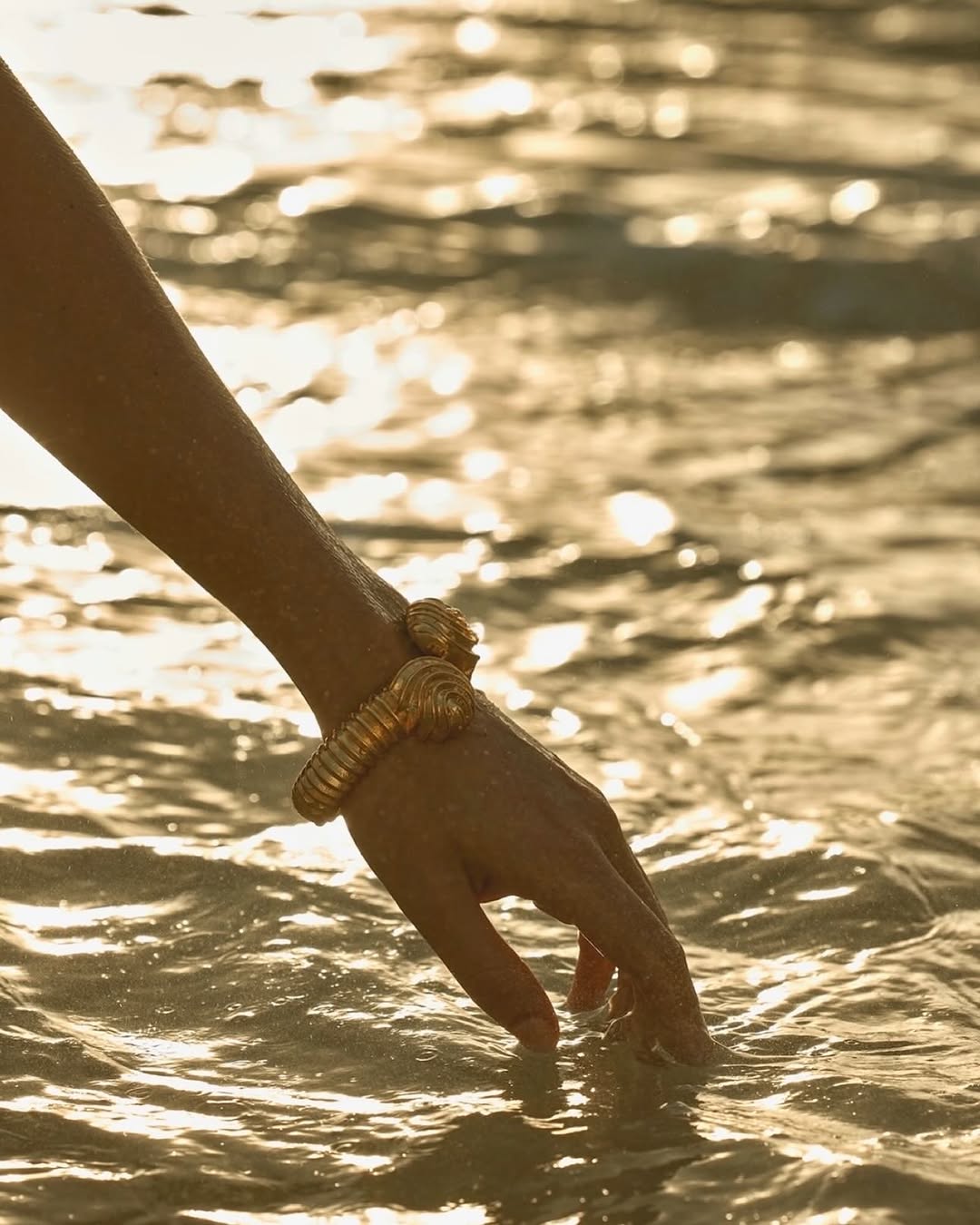 Close-up of a woman's hand with gold bracelet touching the water; the light of the sun reflects off the water on the shore.