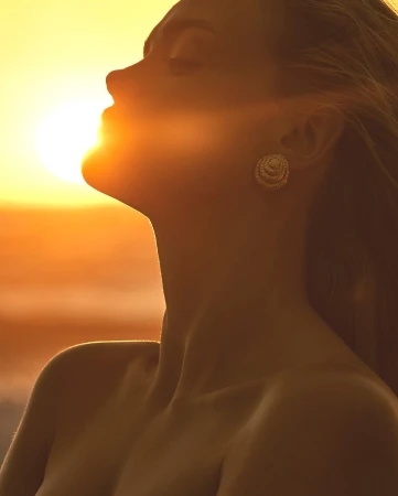 Close-up of a woman with flowing hair silhouetted against a vibrant sunset, highlighting skin tones and jewelry. Soft, golden lighting.