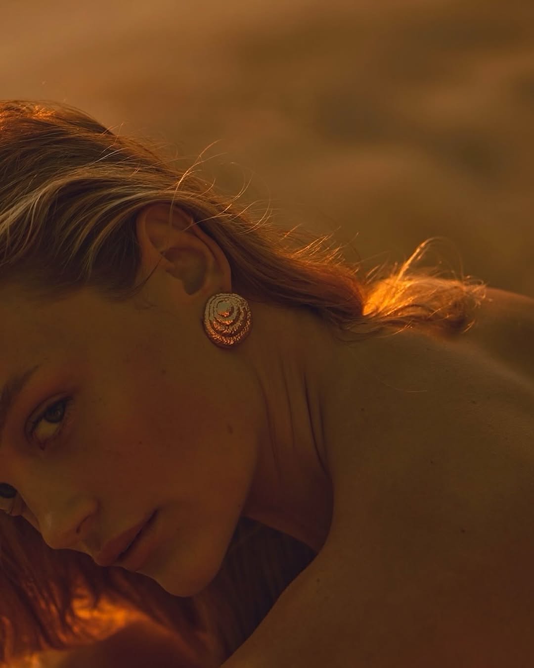 Close-up shot of a blonde woman with golden round earrings bathed in warm, golden light in an indoor environment, beauty shot.