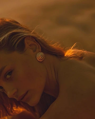Close-up shot of a blonde woman with golden round earrings bathed in warm, golden light in an indoor environment, beauty shot.