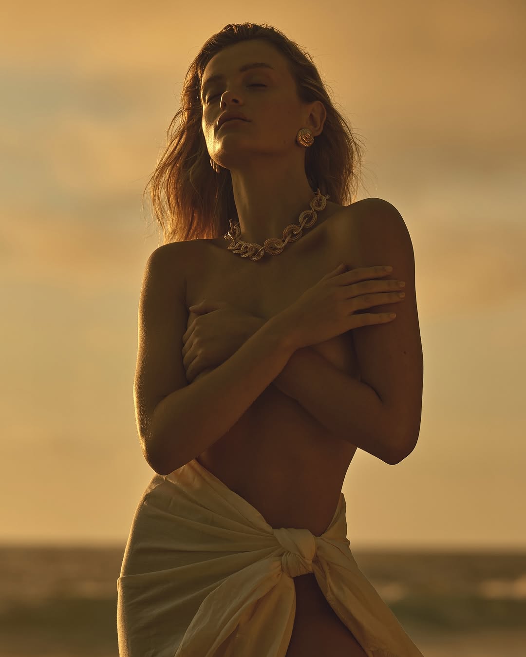 A tanned woman stands outside at the beach wearing jewelry and wrapped in white cloth, in front of ocean waves at sunset.