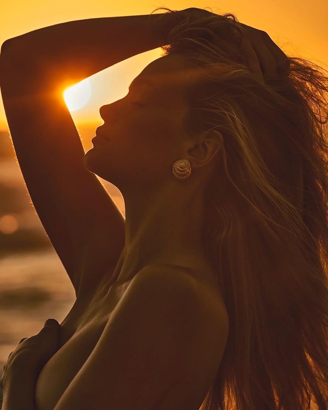 Silhouette of a woman posing at sunset, hand covering breast, long hair, earring detail, against an ocean backdrop during golden hour.