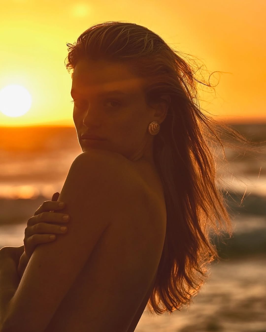 Golden hour portrait of a woman on the beach, facing away from the camera, with ocean waves and a bright sunset in the background.