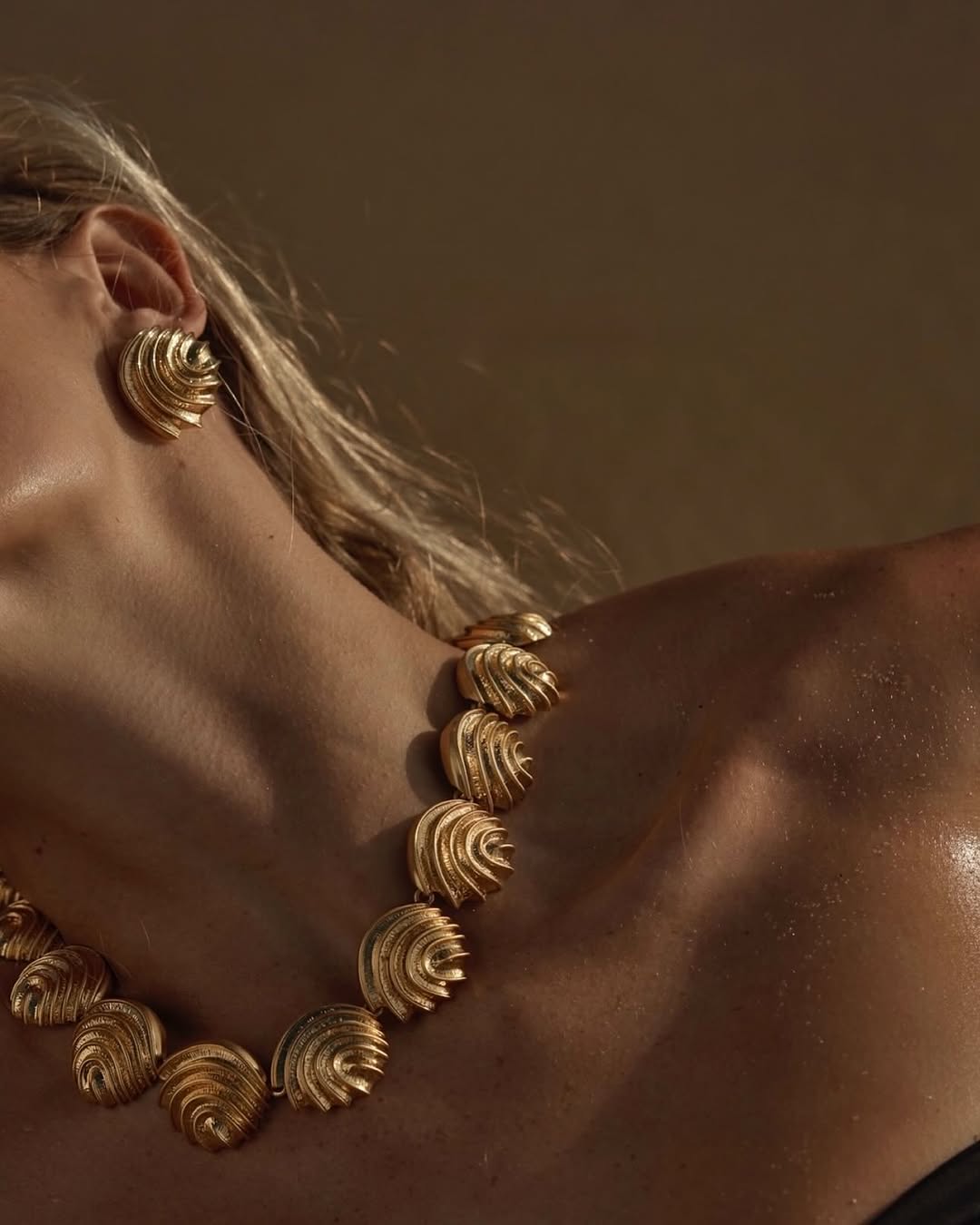 Close-up of a blonde woman wearing a gold necklace and earrings with a spiral design, highlighting jewellery and skin texture in soft light.