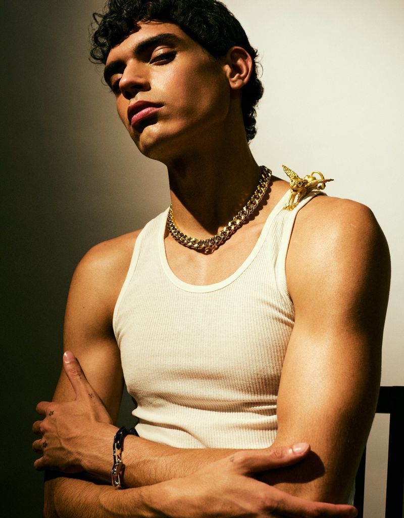 Portrait of a young adult man wearing a tank top and gold chain necklace, posed indoors with studio lighting and a neutral background.