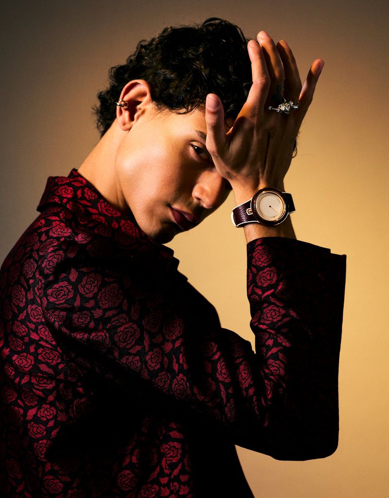 Portrait of a young adult with curly dark hair wearing a rose-patterned jacket, posed in front of a muted brown-toned background.