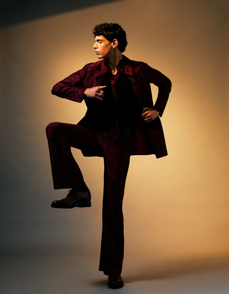 A man in a patterned burgundy suit strikes a stylish pose with one leg raised against a neutral backdrop. The shot is well-lit and emphasizes the texture of the fabric.