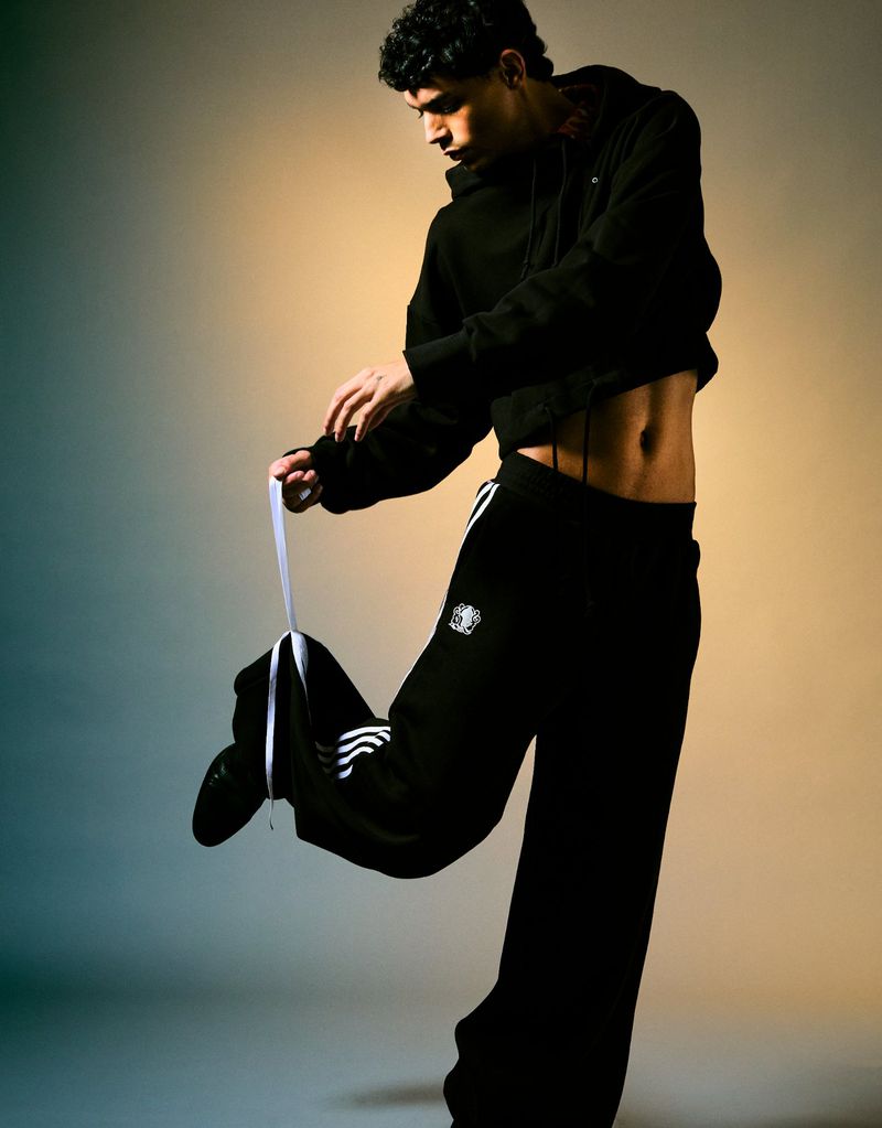 A young adult male strikes a pose indoors wearing a black hoodie and matching track pants with white stripes, under warm studio lighting.