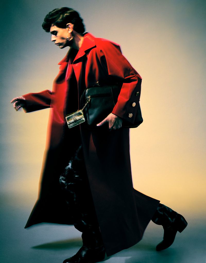 Fashion editorial shot of a person in a red coat walking with a black purse and a smaller metallic bag against a gradient background.