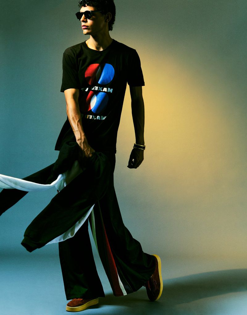 Young adult man in studio wearing sunglasses, patterned t-shirt, track pants, and sneakers in a full length fashion pose.