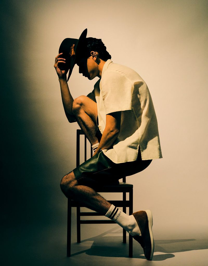 Man sits on a chair in a photo studio with a hat obscuring his face, short sleeves, and shorts, set against a gradient neutral background.