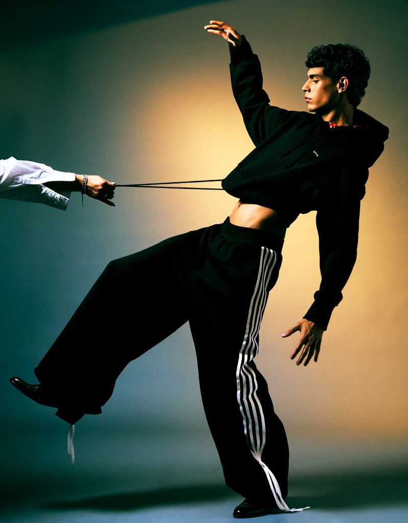Athletic young man in black sportswear posing dynamically in studio setting, pulled by elastic band. Fashion editorial photography.