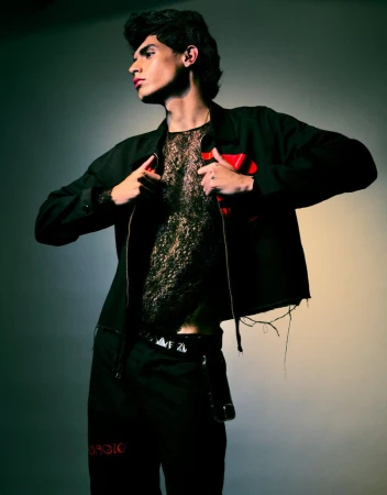 A stylish young man in a black jacket and lace top poses indoors against a gradient background in a fashion-forward editorial shot.