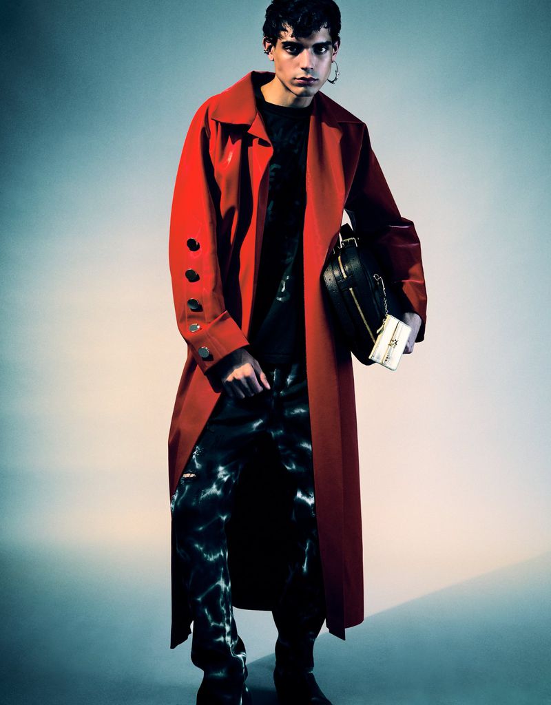 Fashion shot of a young adult man with a black shirt, patterned jeans, and a red trench coat, holding a black purse in a studio setting.
