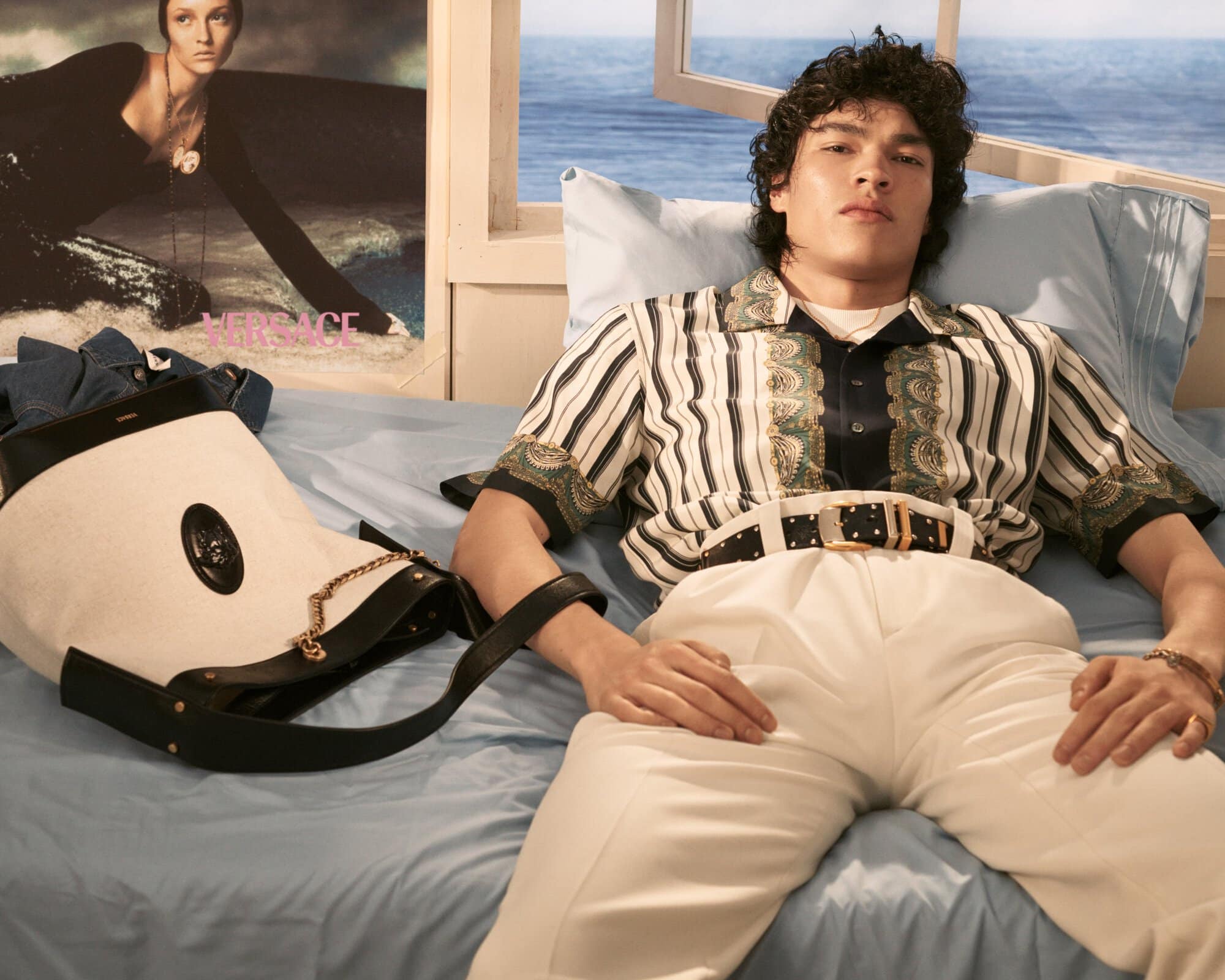 A young male model lying on a bed wearing a patterned short-sleeve shirt and white trousers with a black bucket bag on the sheets.