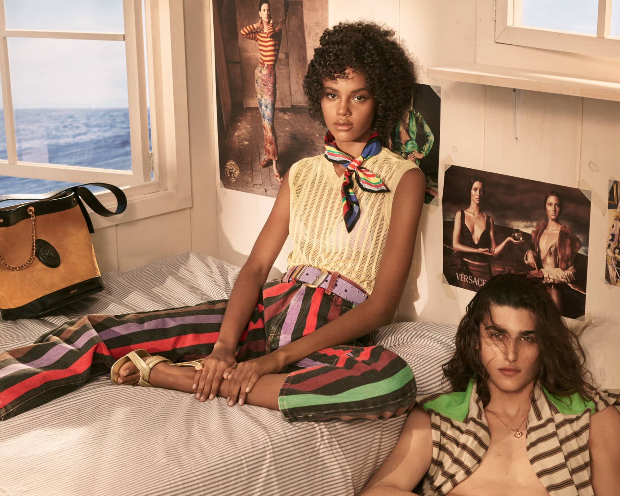 Model with dark curly hair wearing yellow top and striped trousers sitting on a bed beside a man