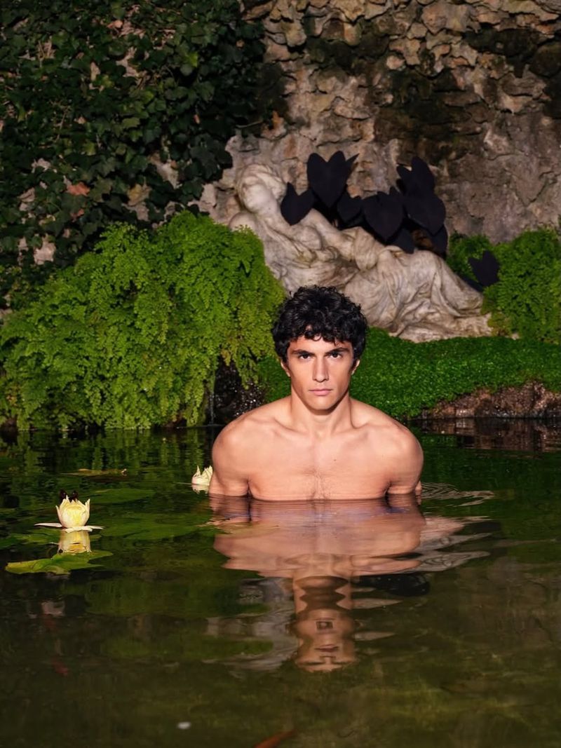 A man submerged up to his chest in dark water with lily pads in front of a stone grotto with statues and lush green plants.
