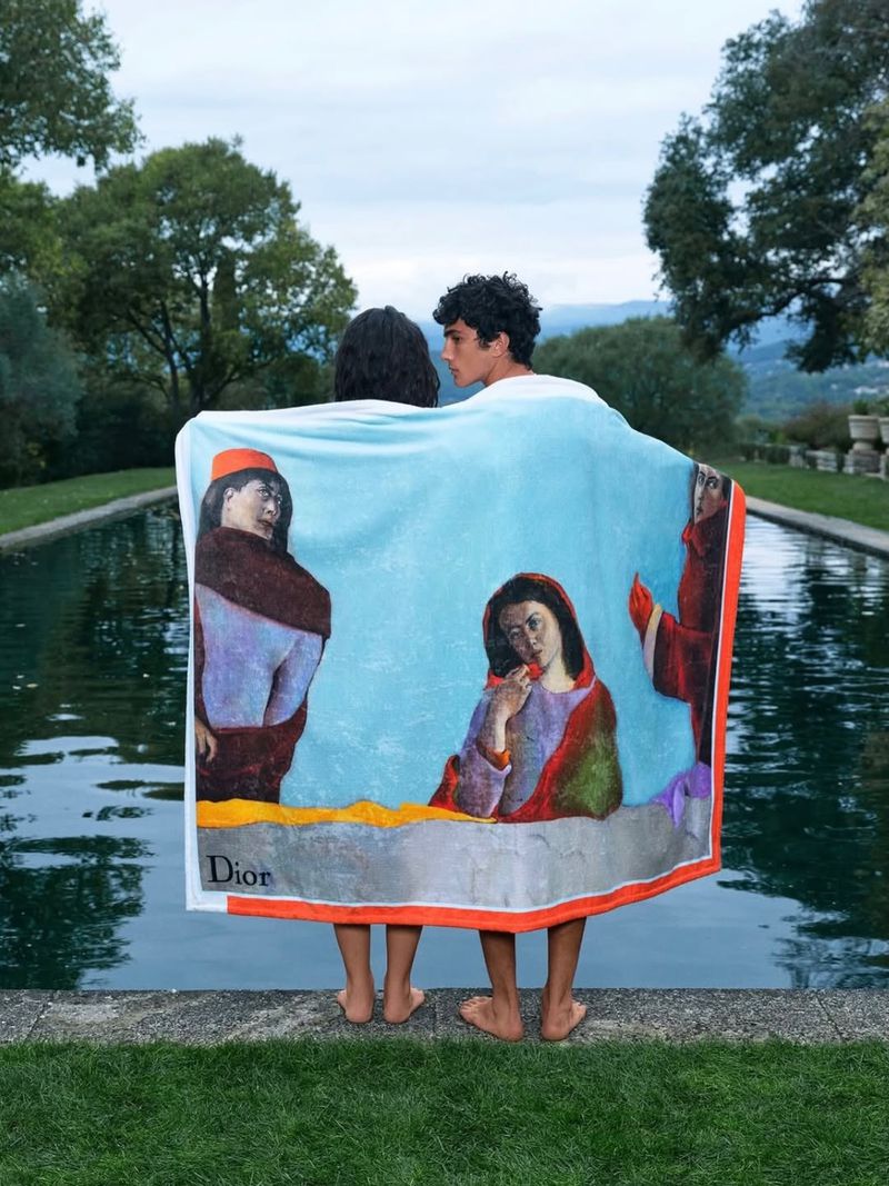 Two models wrapped in a decorative Dior beach towel standing by a tranquil outdoor pool surrounded by greenery.