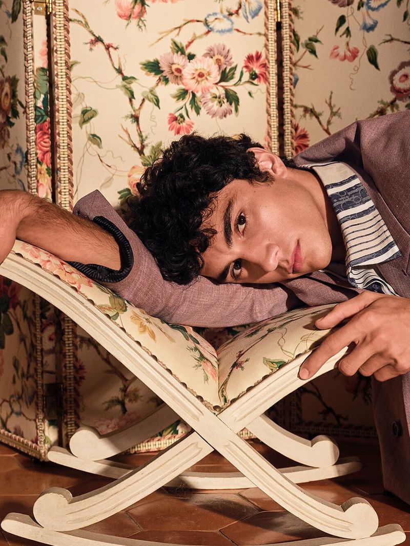 Model resting arm on an ornate folding stool against floral patterned wallpaper in Dior Men campaign