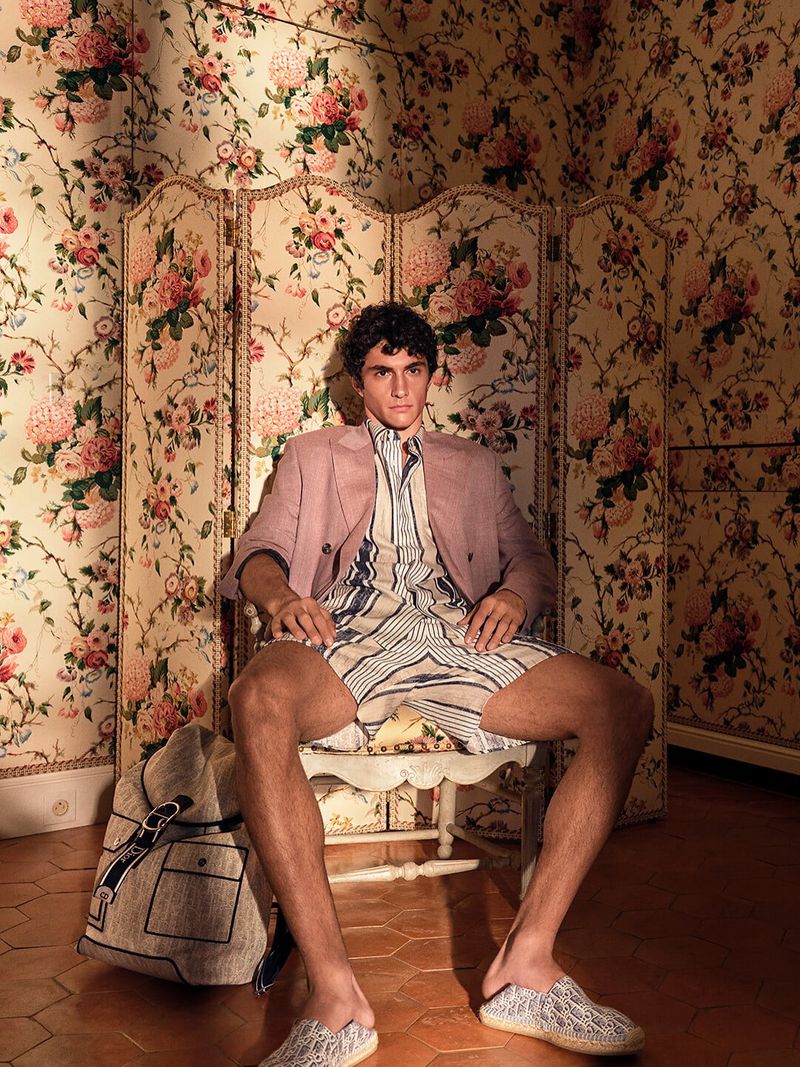 Model seated on a chair in a floral-patterned room wearing a pink blazer and patterned shorts for Dior Men