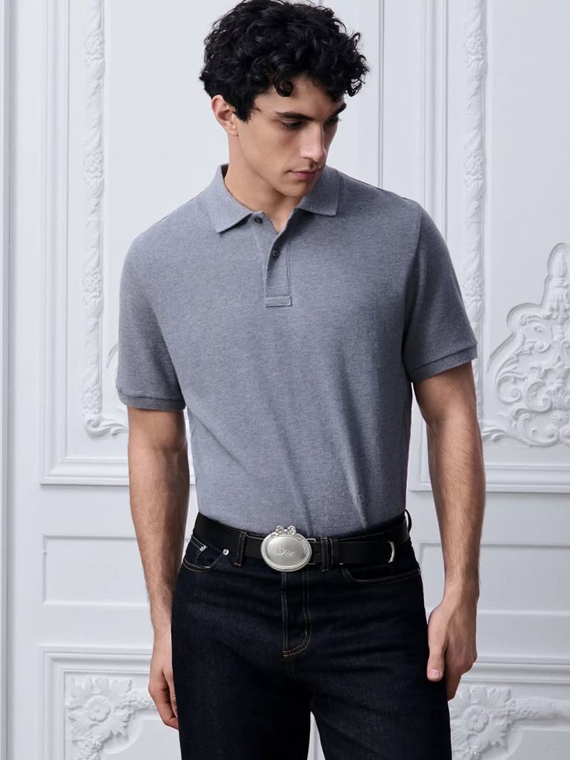 Model wearing a grey polo shirt and black jeans with an ornate belt against an elaborate white paneled wall