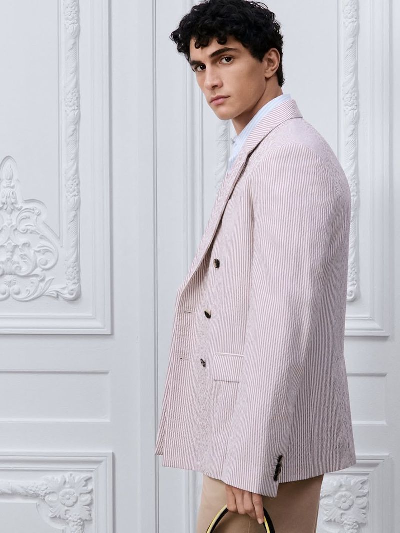 Male model wearing a pink and white striped double-breasted blazer against an ornate white neoclassical wall panel background