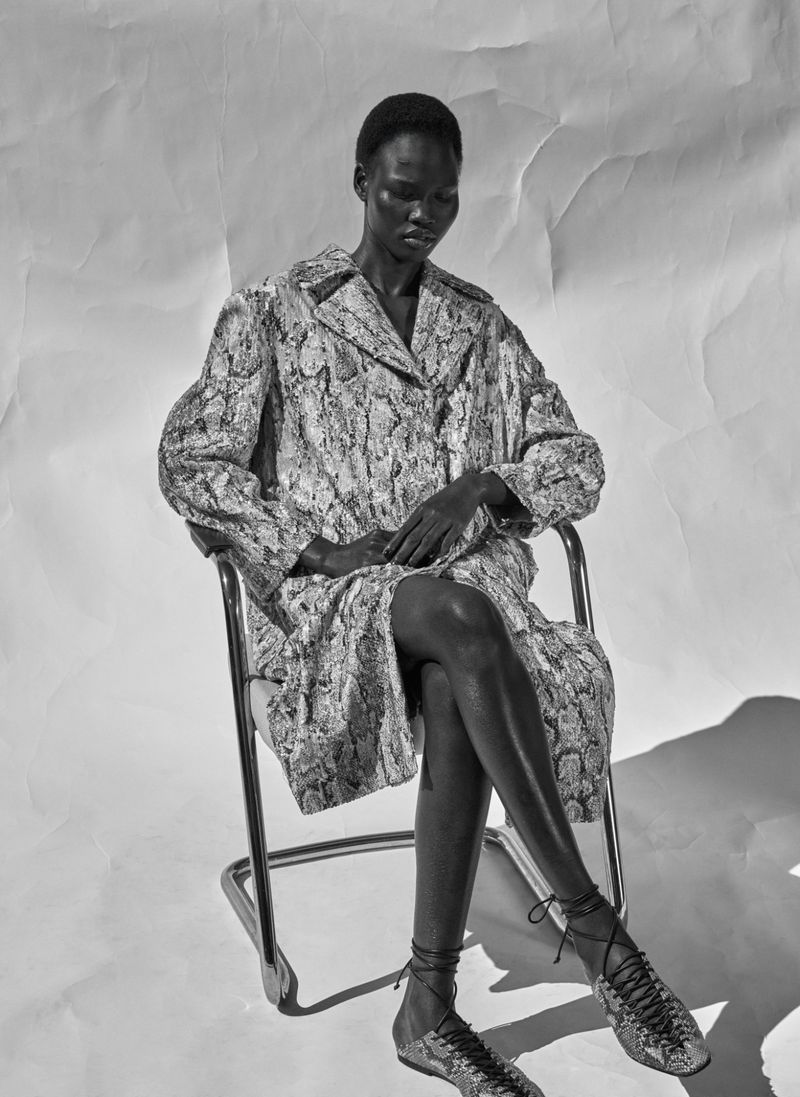 Black and white shot of a Black female model in a patterned coat sitting in a chair. The background is a creased white paper.
