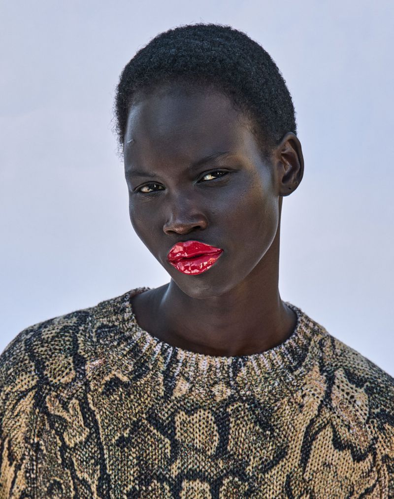 Close-up studio shot of a dark-skinned woman with short black hair wearing a patterned sweater and glossy red lipstick, neutral background.