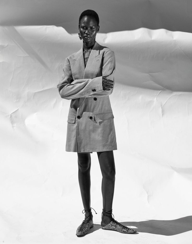 Black and white fashion shot of a Black female adult model wearing a blazer dress and lace-up shoes against a white backdrop.