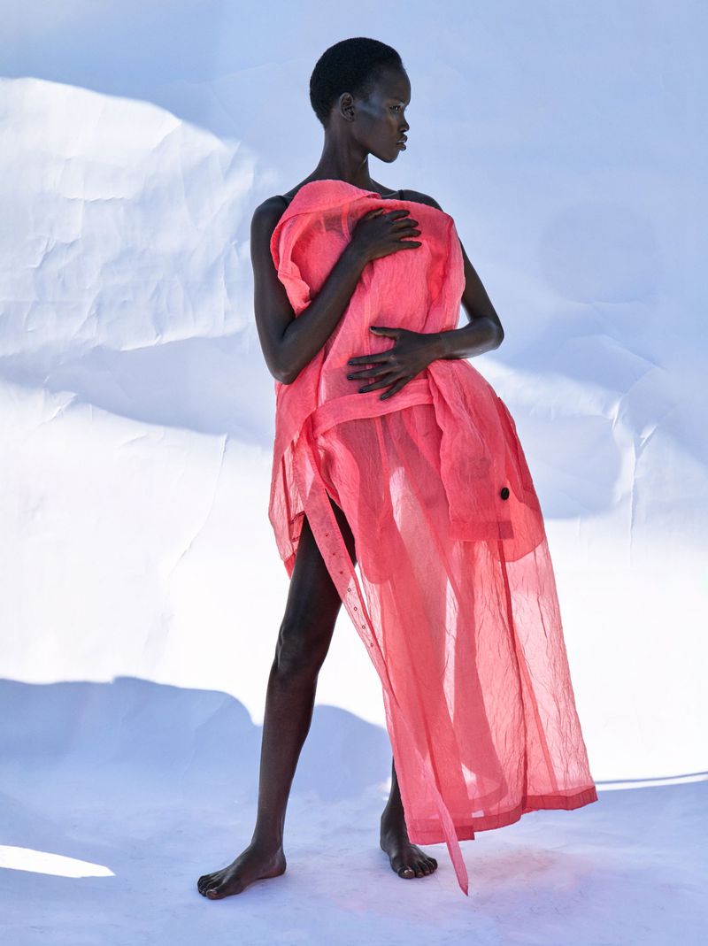An African model poses in a translucent coral dress against a white backdrop in a fashion editorial style image. She gazes off to the right.