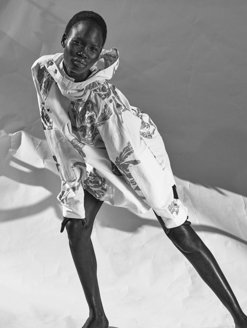 Monochrome portrait of a slender African woman in an ornate hooded shirt, angled pose against a textured paper backdrop in soft lighting.