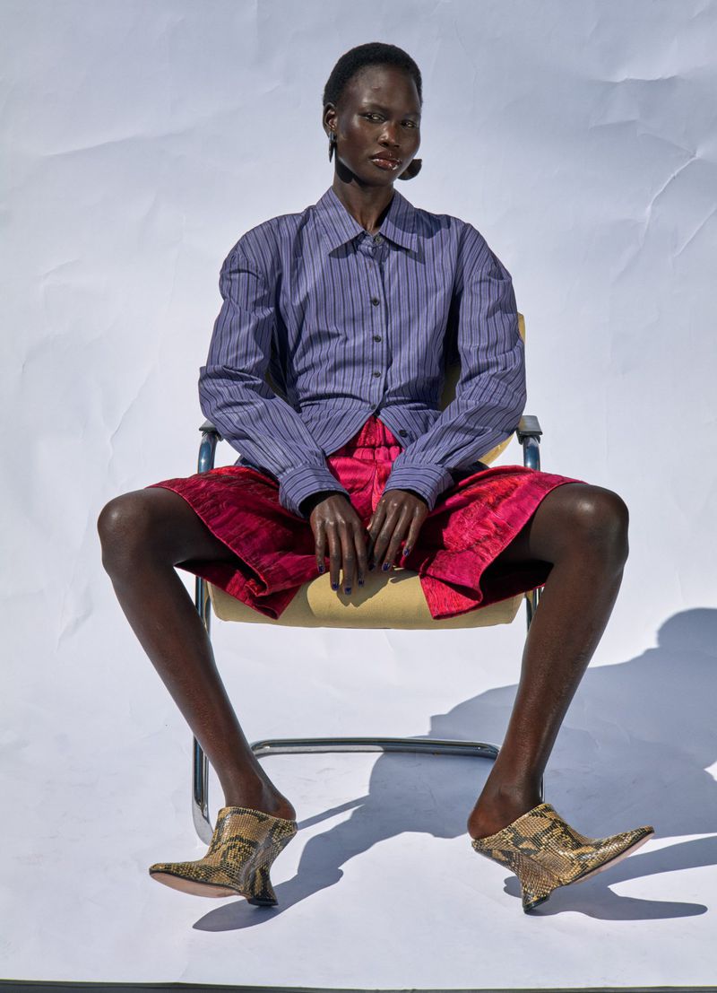 Fashion shot of an African-descent female model in a blue button-up shirt and red shorts, seated on a chair in front of a white backdrop.