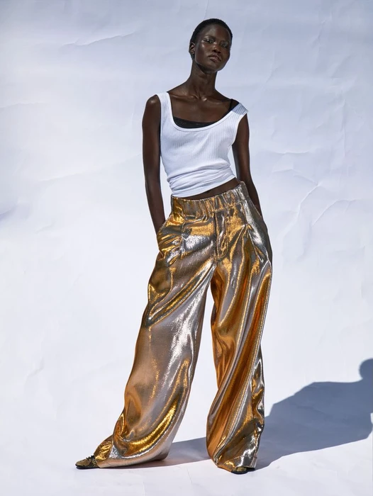 Model Chudier Billy wearing a white tank top and metallic gold wide-leg trousers against a white seamless background