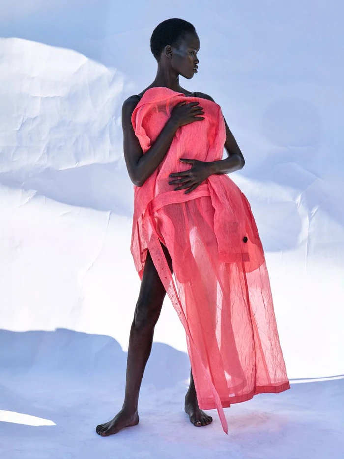 Model Chudier Billy wrapped in a sheer pink garment standing barefoot against a bright white paper backdrop.