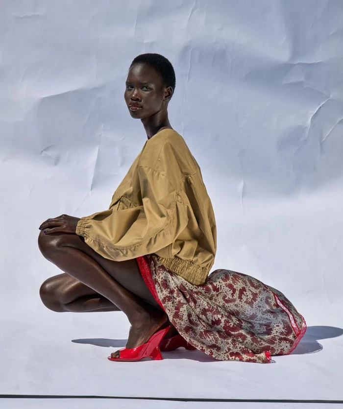 Model Chudier Billy crouching in a tan jacket, printed skirt, and red heels against a crinkled white backdrop.