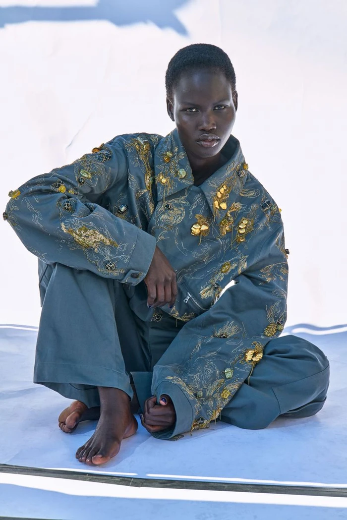 Model Chudier Billy seated barefoot wearing an embellished teal jacket and matching trousers on white seamless background.