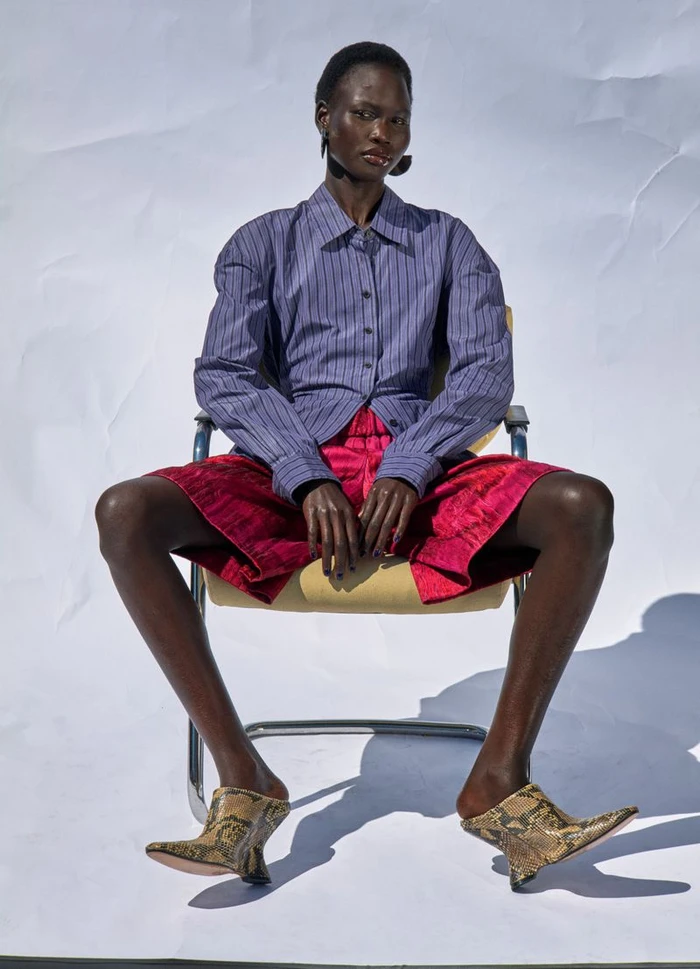Model Chudier Billy seated in a chair wearing a striped shirt, red shorts, and snakeskin mules for Twin Magazine.
