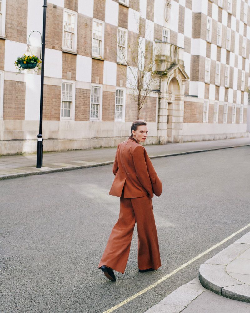 Model in rust-coloured blazer and trousers walking on an urban street with a patterned building exterior in the background
